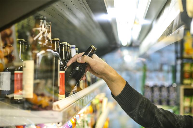 A bottle of beer is picked up by a person’s hand from a store shelf that contains multiple other bottles.