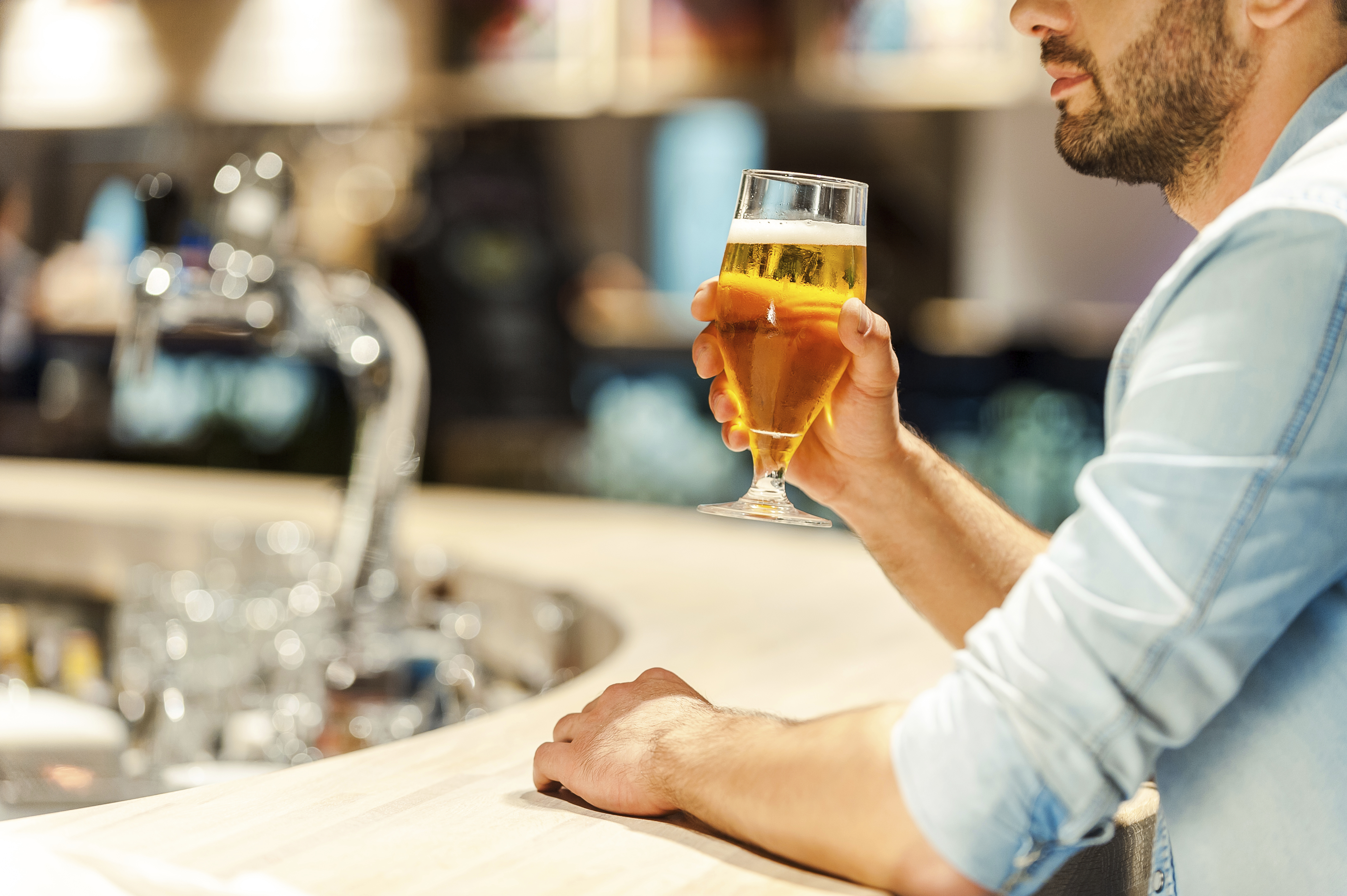 Homme avec un verre de bière à la main, assis au comptoir d’un bar avec des bouteilles et des fûts flous en arrière-plan.
