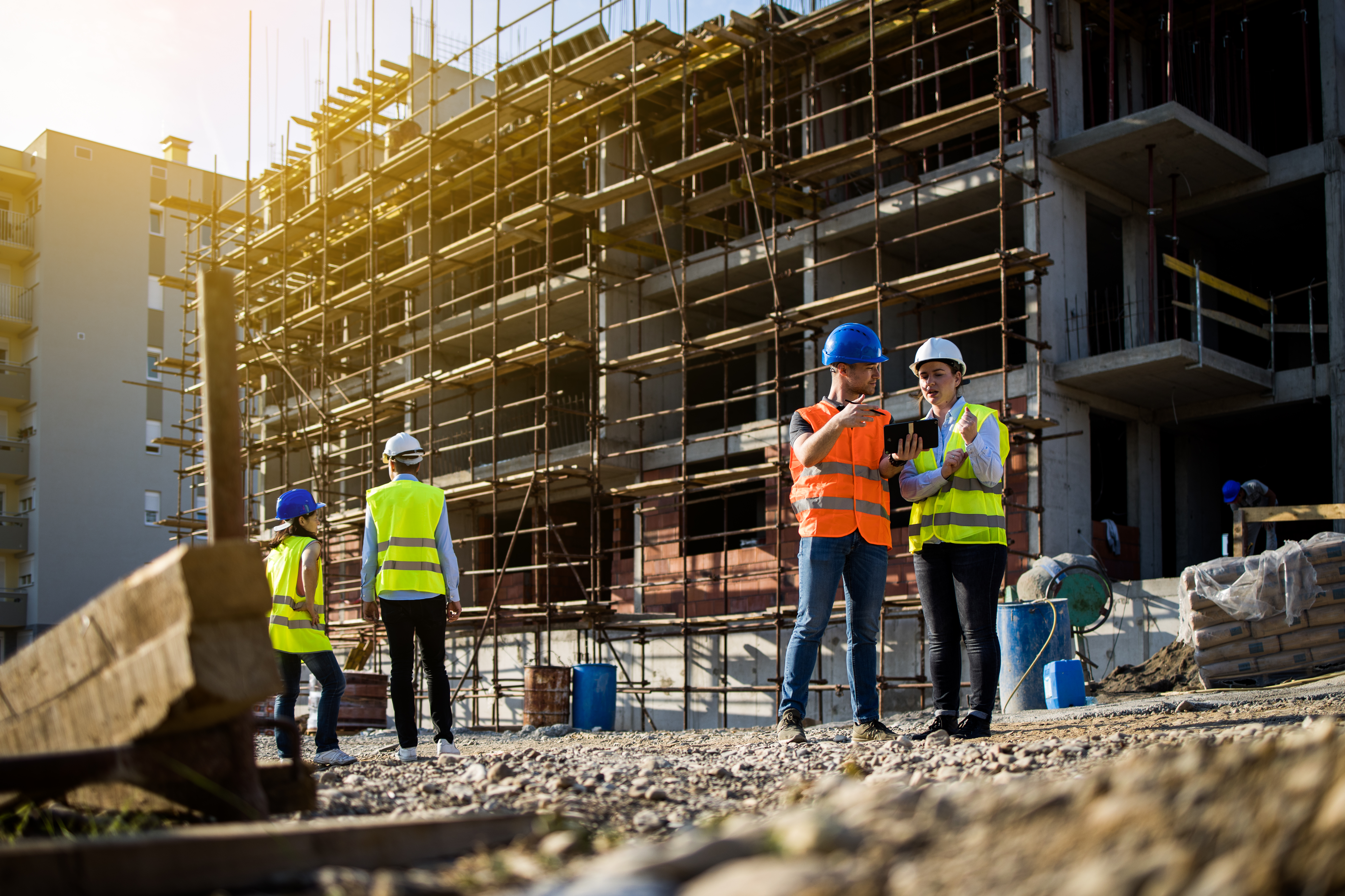 Construction workers in safety gear discussing plans at a building site with scaffolding in the background during sunset.