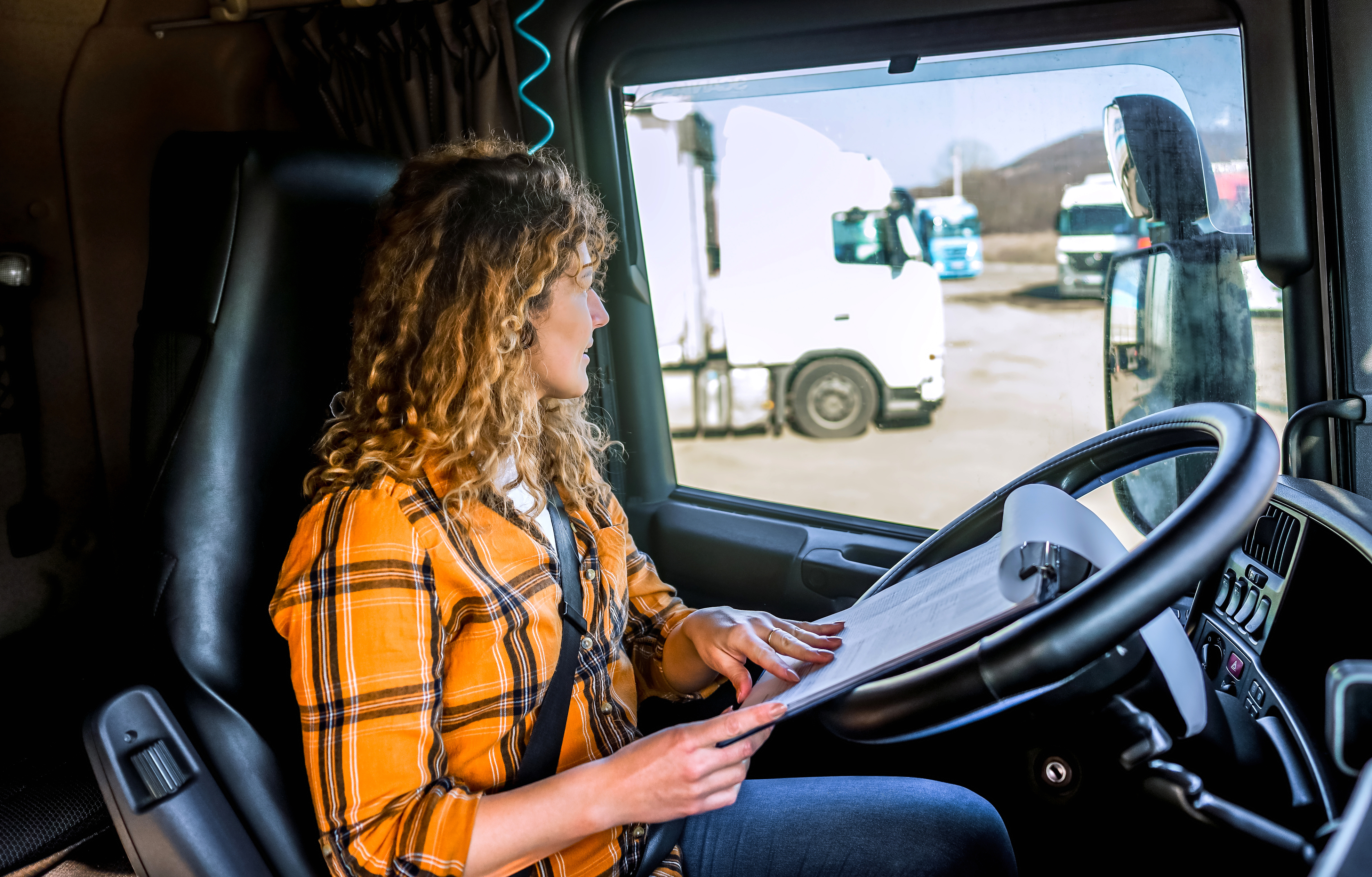 Woman in a yellow plaid shirt sitting in a truck cabin reading documents on a clipboard that is resting on the steering wheel.