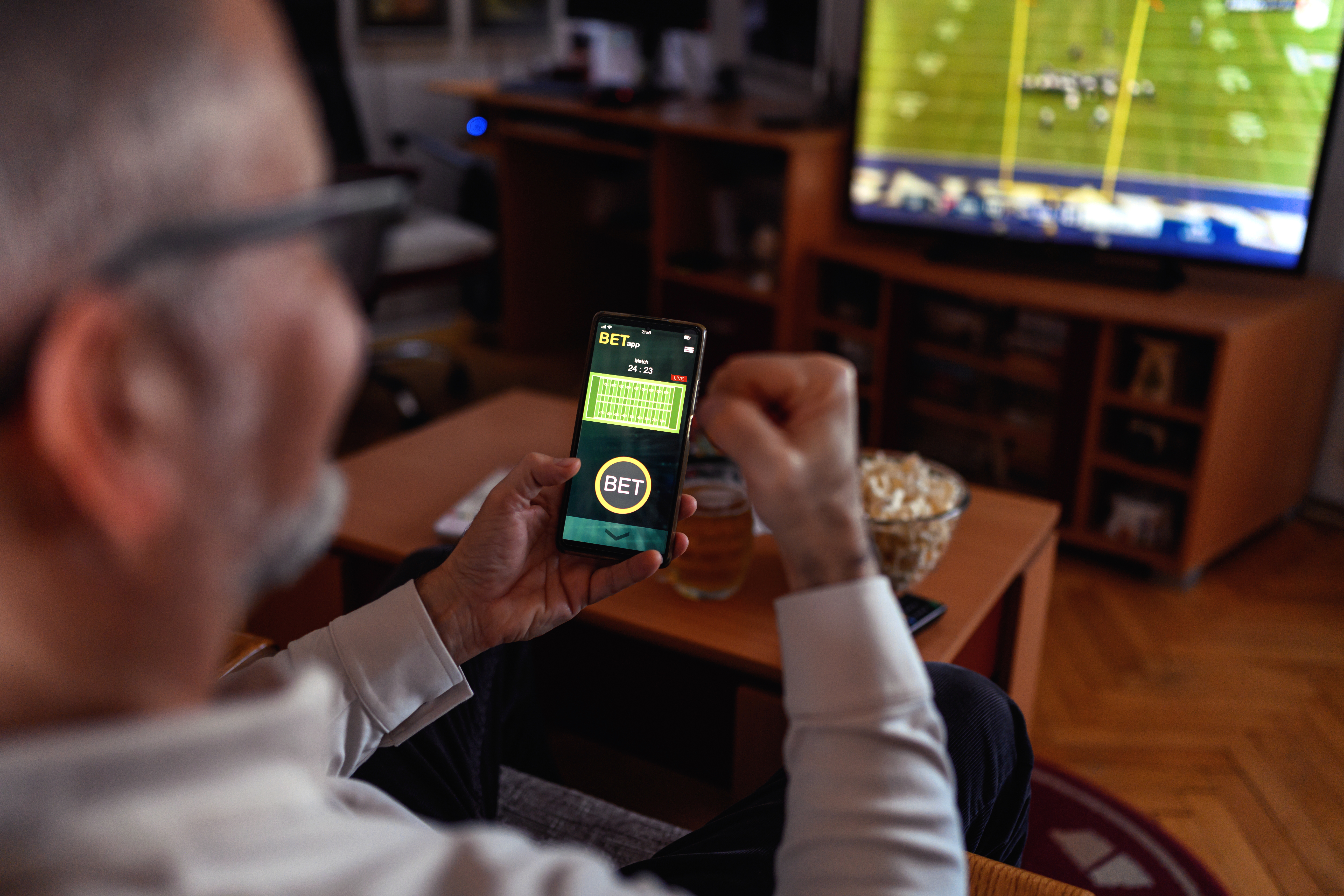 Man holding smartphone with a betting app open while watching a football game on TV in a cozy living room.