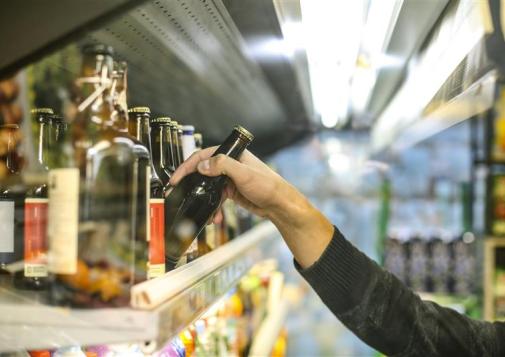A bottle of beer is picked up by a person’s hand from a store shelf that contains multiple other bottles.