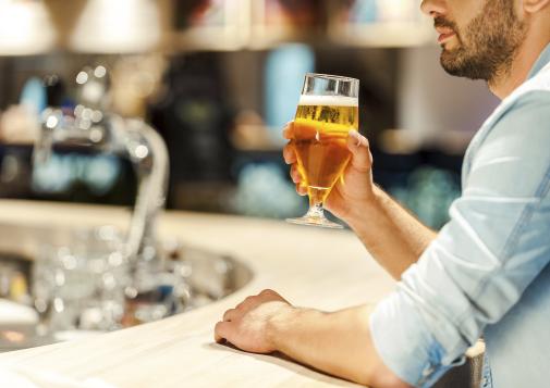 Man holding a glass of beer while sitting at a bar counter with a blurred background of bottles and taps.