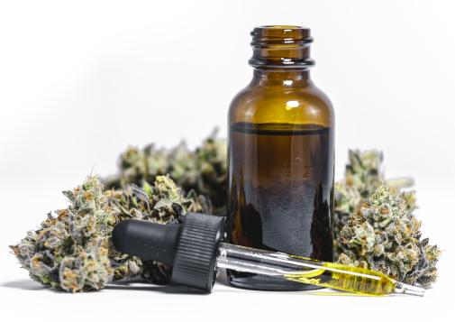 Amber glass bottle with a dropper filled with cannabis oil surrounded by dried cannabis buds on a white background.