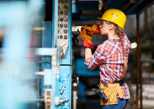 Female factory worker wearing a yellow hard hat and plaid shirt operating machinery in an industrial setting.