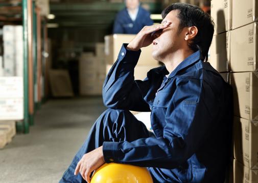Male warehouse worker in blue uniform sitting on the floor, resting against stacked boxes, holding a yellow hard hat and wiping sweat from his forehead.