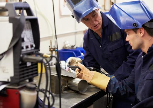 Two workers in protective gear welding a metal pipe in an industrial workshop setting.