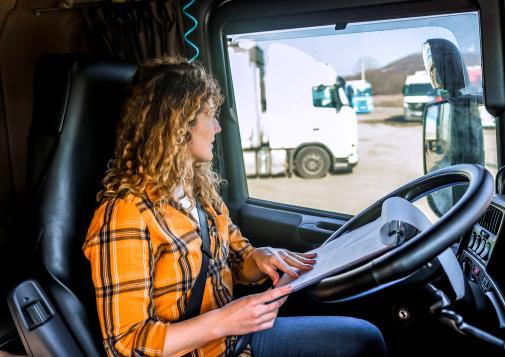Woman in a yellow plaid shirt sitting in a truck cabin reading documents on a clipboard that is resting on the steering wheel.