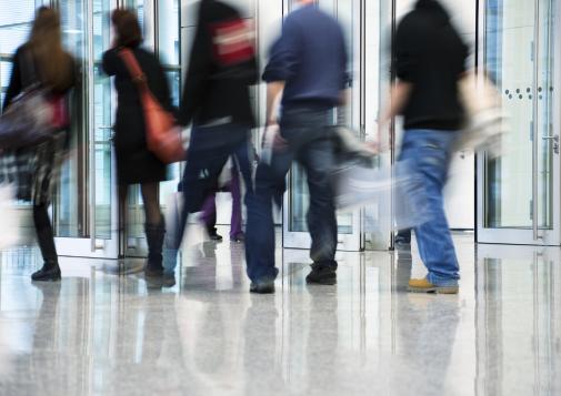 Blurred motion of people walking through a modern glass entrance in a busy urban building lobby.