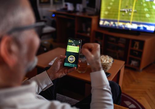 Man holding smartphone with a betting app open while watching a football game on TV in a cozy living room.