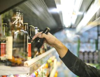 A bottle of beer is picked up by a person’s hand from a store shelf that contains multiple other bottles.