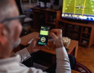 Man holding smartphone with a betting app open while watching a football game on TV in a cozy living room.