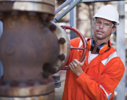 Male industrial worker in orange safety gear and white helmet turning a large red valve on a rusted pipe system outdoors.