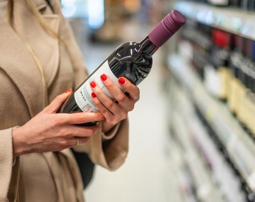 Person wearing a beige coat holding a bottle of red wine while shopping in a liquor store aisle.