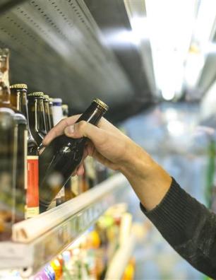A bottle of beer is picked up by a person’s hand from a store shelf that contains multiple other bottles.