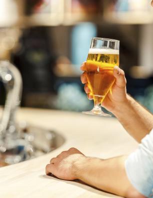 Man holding a glass of beer while sitting at a bar counter with a blurred background of bottles and taps.