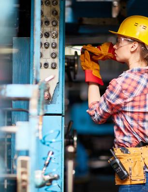 Female factory worker wearing a yellow hard hat and plaid shirt operating machinery in an industrial setting.