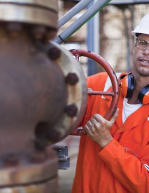 Travailleur industriel portant des vêtements de sécurité orange et un casque de protection blanc qui fait tourner une grande valve rouge sur un tuyau rouillé, à l’extérieur.