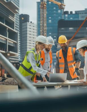 Construction workers in safety gear reviewing blueprints on an urban building site with cranes and skyscrapers in the background.