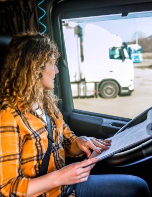 Woman in a yellow plaid shirt sitting in a truck cabin reading documents on a clipboard that is resting on the steering wheel.