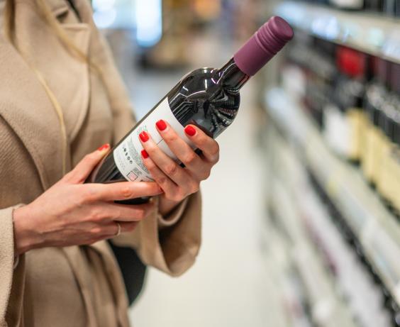 Person wearing a beige coat holding a bottle of red wine while shopping in a liquor store aisle.
