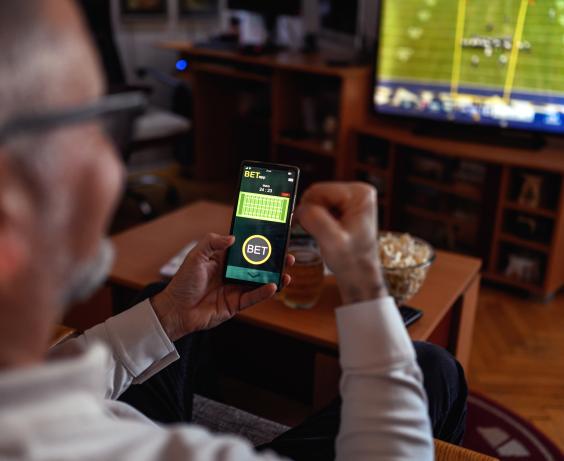 Man holding smartphone with a betting app open while watching a football game on TV in a cozy living room.